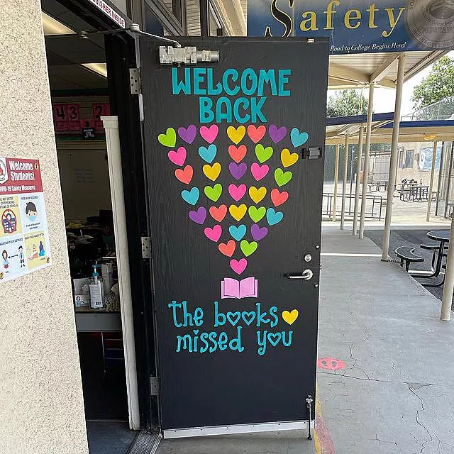 A door says welcome back the books missed you. There is an open book and a lot of rainbow hearts above it.