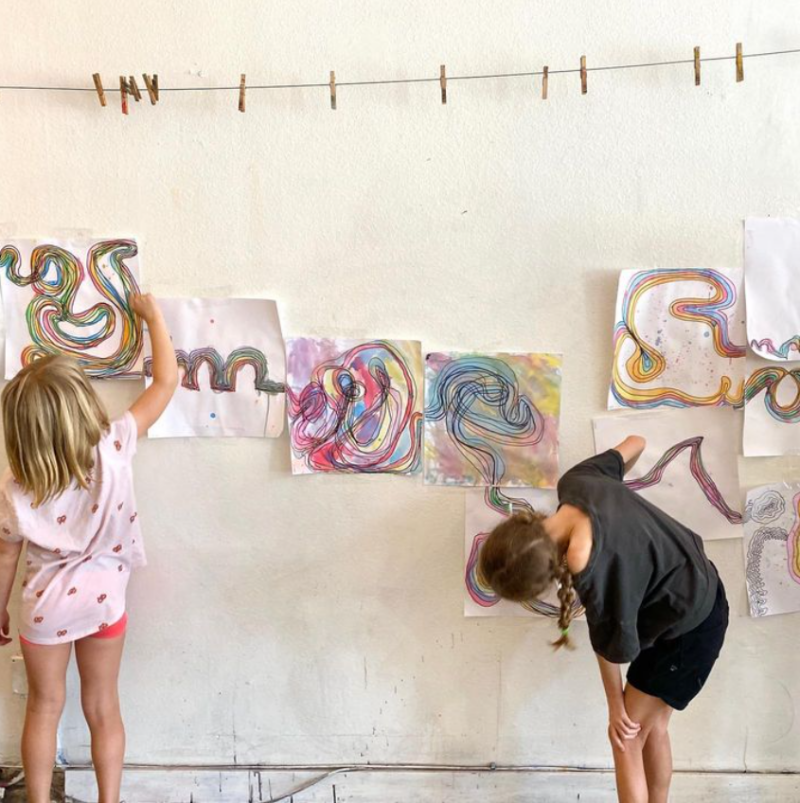 Two children stand in front of a wall that has a bunch of swirly drawings lined up horizontally on it.
