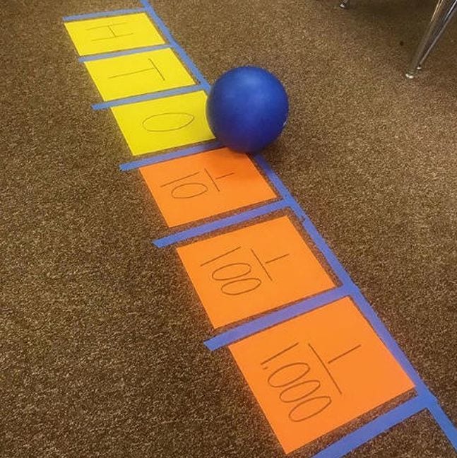 Large number line made of colorful paper squares and blue tape, with a rubber ball marking a decimal