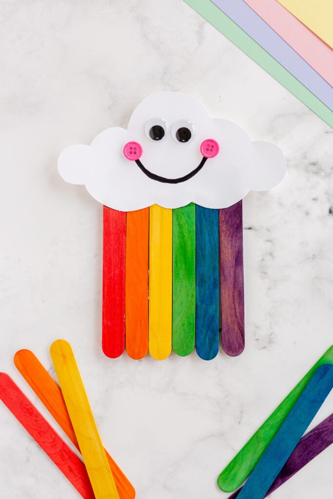 A white cloud has a smiley face on it with rainbow colored popsicle sticks coming out of it.