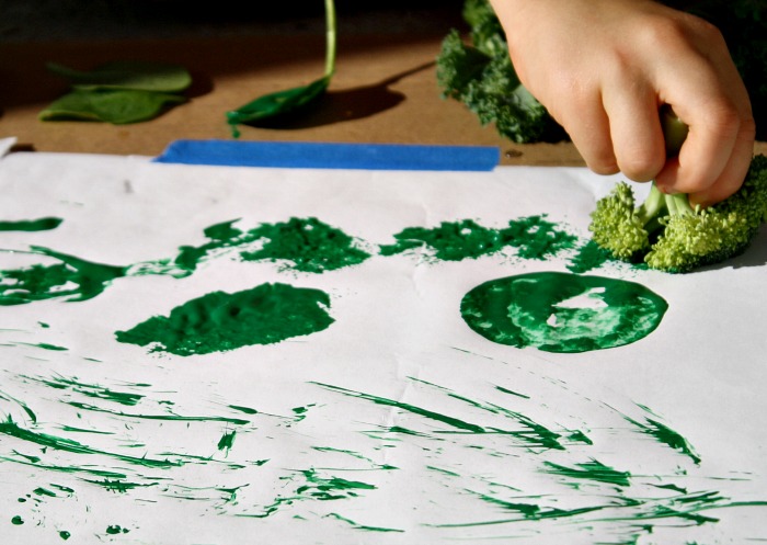 A white piece of paper is seen with green paint splotches on it. A hand is seen holding a piece of broccoli to the paper.