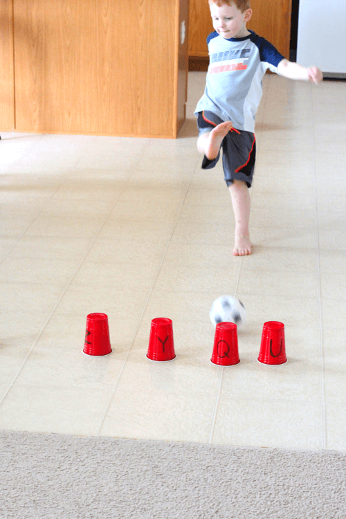 a child kicking a ball at a row of cups with letters on them for a preschool activity