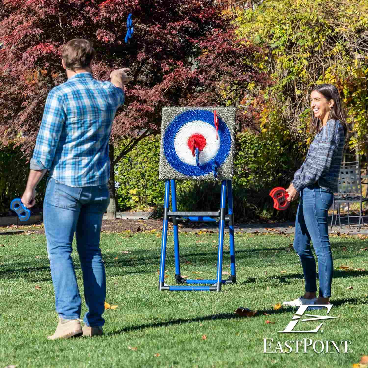 Two people throwing toy axes at a bristle-covered target on a blue metal frame