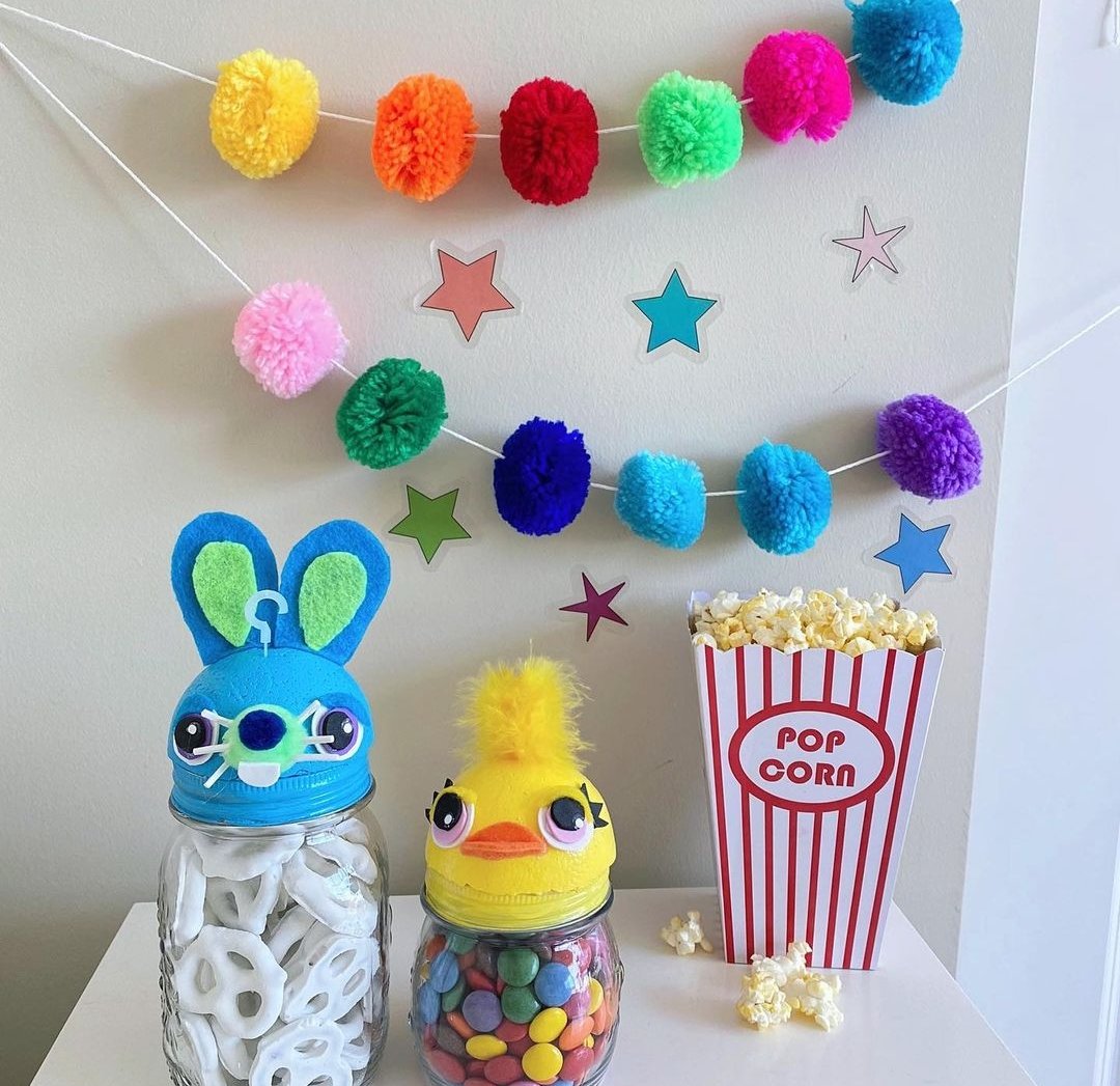 Two containers topped with animal heads, filled with snacks, against a pom pom garland background with a container of popcorn