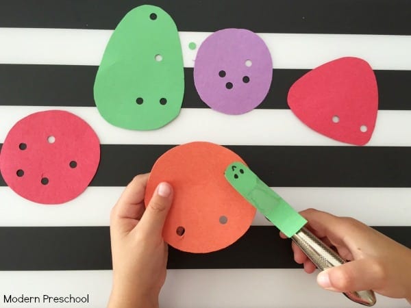 A child's hands punch holes into construction paper shapes on a background of black and white stripes