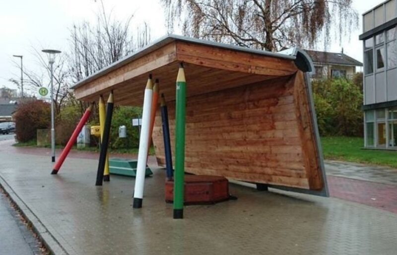 A covered school bus stop has creative columns made to look like colored pencils and a cover that looks like an open book