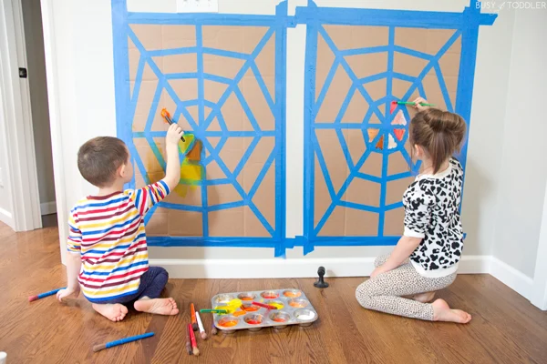 Two children painting spiderwebs taped to the wall