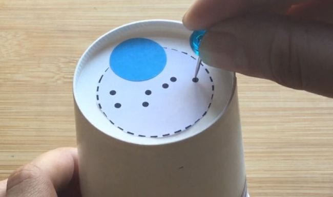 DIY star projector for a second grade science experiment, constructed of an upside-down paper cup with seven holes punched on the bottom of it, a blue circle the size of a quarter, and a bigger white circle surrounded by dashes.